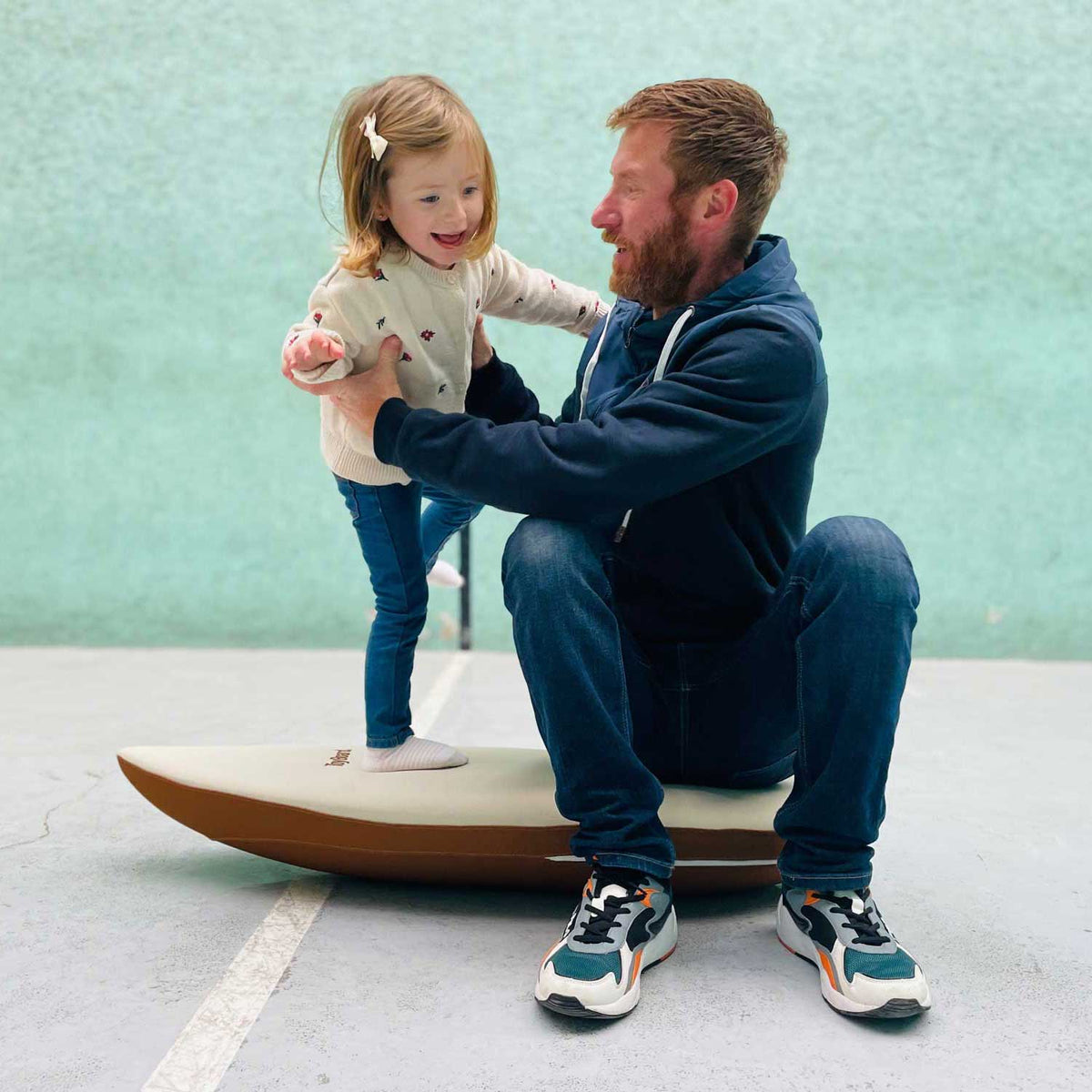 Un adulte assis sur une planche au sol soutient un enfant debout à ses côtés.